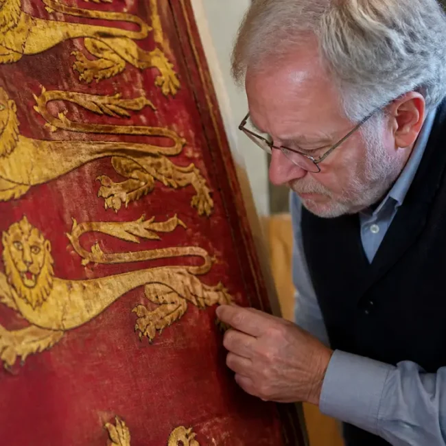 Gros plan sur un artisan examinant une tapisserie médiévale pour comprendre pourquoi le blason Normandie pourquoi utilisé s'inspire des Plantagenêt.