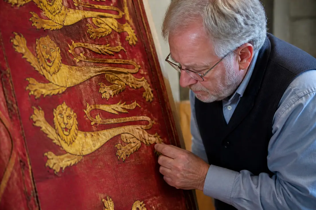Gros plan sur un artisan examinant une tapisserie médiévale pour comprendre pourquoi le blason Normandie pourquoi utilisé s'inspire des Plantagenêt.